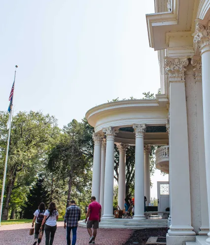 A group of people walk towards a grand building with tall white columns and elaborate architectural details. The setting is outdoors, with trees and an American flag on the left, giving a historical, serene atmosphere.