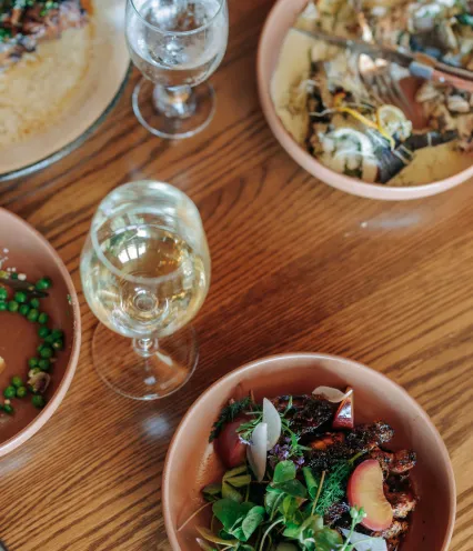 A wooden table set with plates of gourmet food, including a salad with fresh greens and plums, and a dish with peas. Two glasses of white wine add elegance.