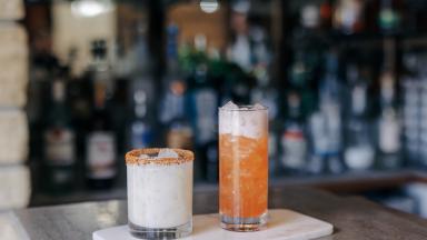 Two refreshing cocktails on a marble tray: a creamy white drink with a rim of spices and a vibrant orange drink with ice.