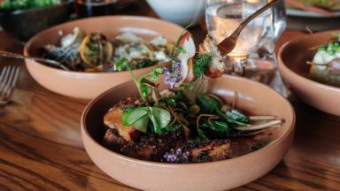 A gourmet dish featuring succulent meat topped with herbs and edible flowers, being lifted by a fork, with additional plates in the background.