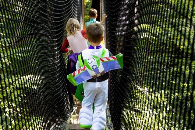 A child in a Buzz Lightyear costume walks on a forest bridge, followed by two other kids, surrounded by lush greenery.