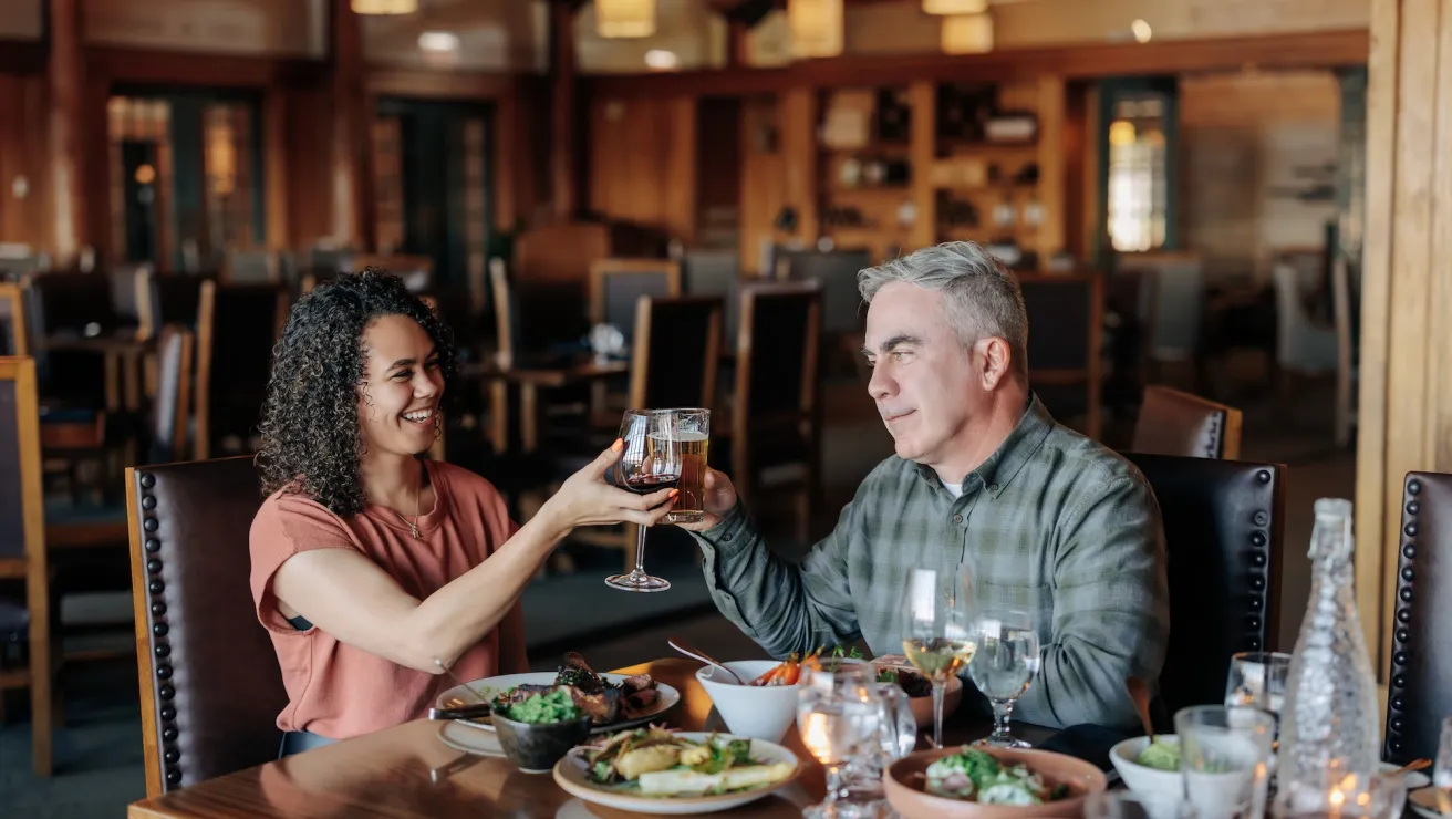 Two people toast with glasses in a cozy restaurant setting, surrounded by plates of food and soft lighting.