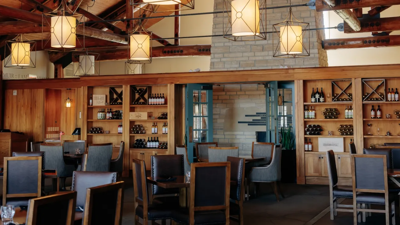 A cozy restaurant interior featuring wooden furniture, decorative light fixtures, and a well-stocked wine display on the back wall.
