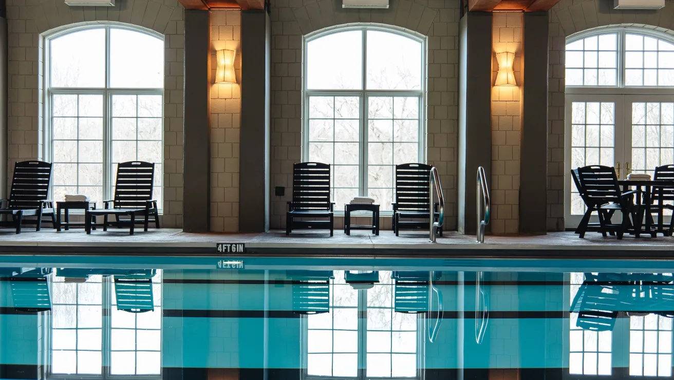 Indoor swimming pool with reflections, featuring lounge chairs, large windows, and warm lighting.