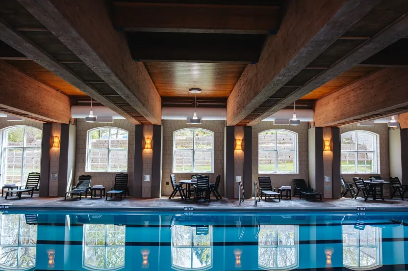 Indoor swimming pool with wooden beams, lounge chairs, and large windows reflecting bright light and greenery.
