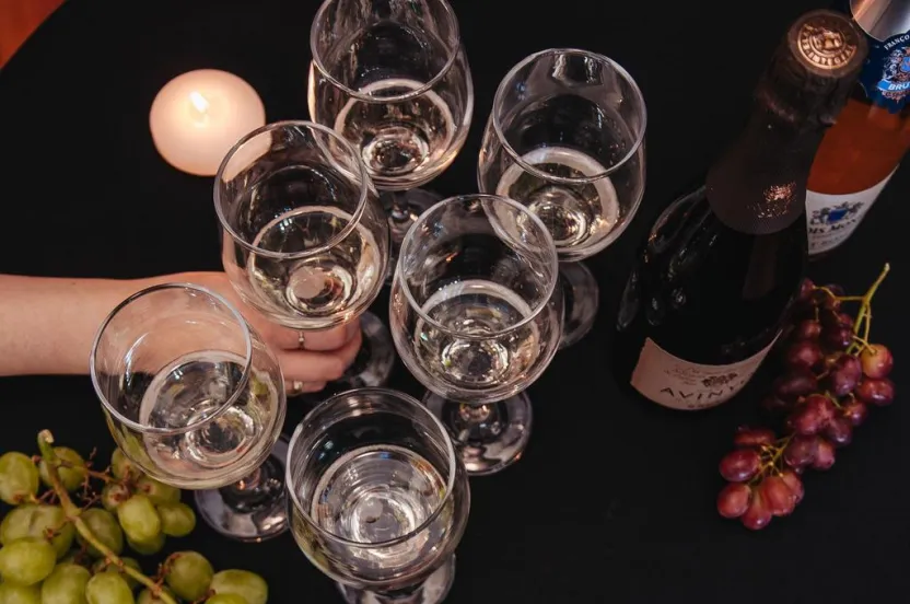 Six wine glasses filled with white wine, a bottle of sparkling wine, grapes, and a lit candle on a black tablecloth.