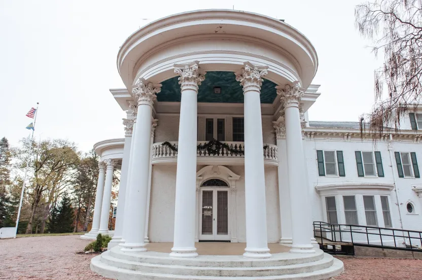 A grand white mansion with classic columns and a circular entrance, decorated with greenery, set against a cloudy sky.
