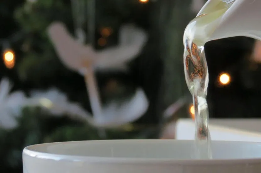 A teapot pours steaming tea into a white cup, set against a softly lit background of a decorated Christmas tree.