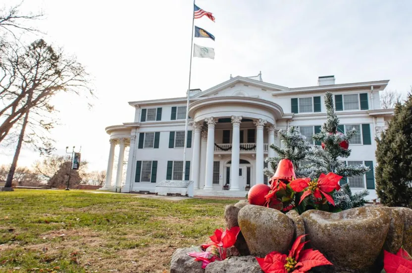 Historic white mansion with green shutters, flags flying, adorned with festive decorations and a small Christmas tree in the foreground.