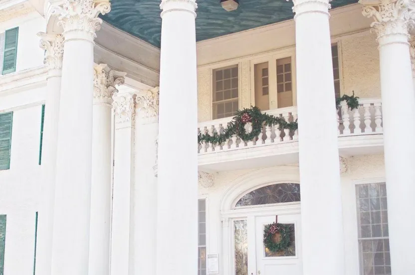 Elegant entrance of a stately building featuring tall white columns, decorative wreaths, and a blue ceiling above the porch.