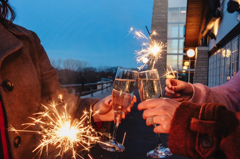 Two people clink champagne glasses, holding sparklers. It's dusk with a festive atmosphere, evoking celebration and joy against a building backdrop.