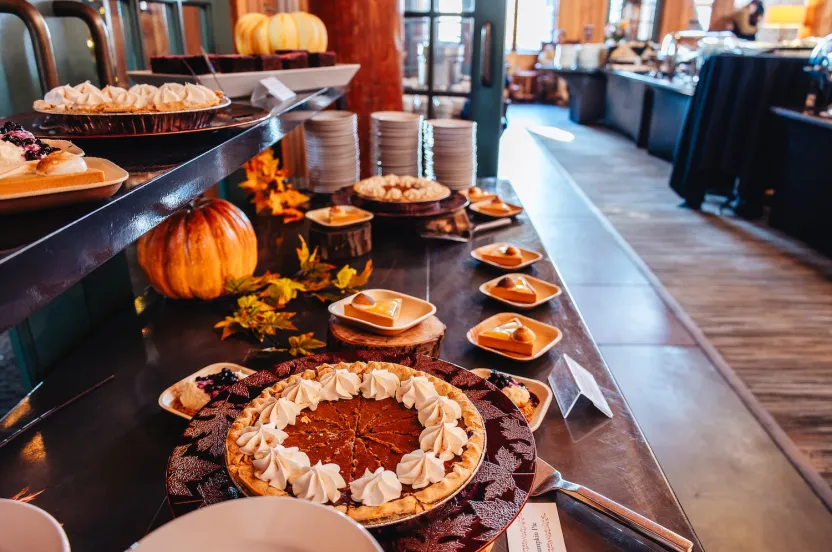 Festive dessert buffet with pumpkin pies topped with whipped cream, surrounded by plates of cheesecake, autumn leaves, and a decorative pumpkin.