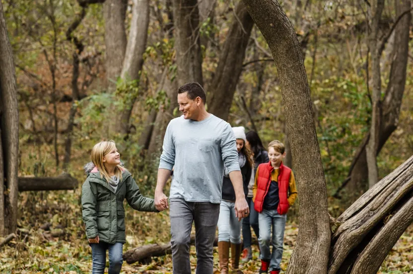 A man and four children stroll through a serene forest, surrounded by autumn foliage and tall trees.