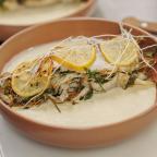 Grilled fish with lemon slices, garnished with herbs and sprouts, resting on a creamy sauce in a rustic bowl.