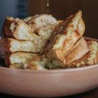 Stacked French toast drizzled with syrup sits on a pink plate, showcasing a deliciously golden and buttery texture.
