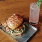 A crispy fried chicken sandwich with a sesame seed bun, topped with slaw, alongside a refreshing pink drink garnished with mint.