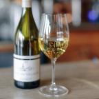 A glass of white wine sits on a marble table beside a bottle labeled "Macon-Villages," with a blurred bar backdrop.