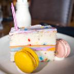A slice of colorful birthday cake with a pink candle, surrounded by yellow and pink macarons on a white plate.